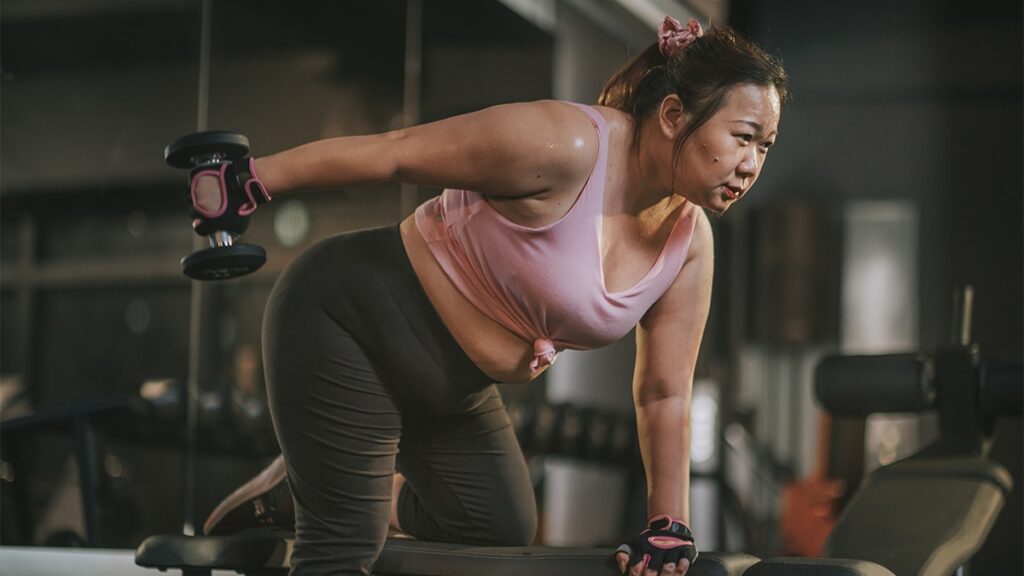 body positive Asian mid adult woman exercising with dumbbells in a lunge position at gym at night